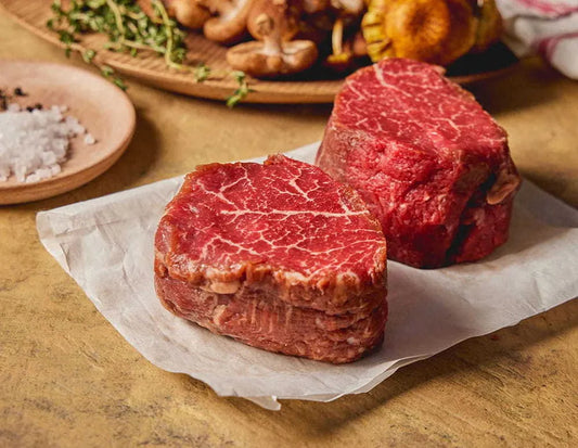 Two raw filet mignon steaks on a piece of parchment paper, with seasonings and other food items in the background.