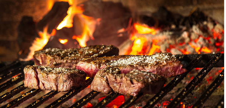 Steaks being grilled over an open flame with glowing coals and embers.