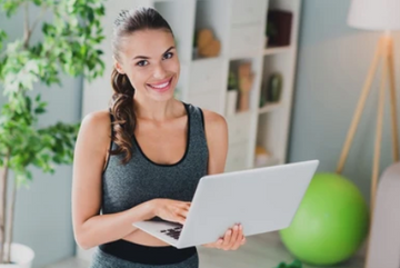woman holding a laptop in athleisure wear with a yoga ball in the background.