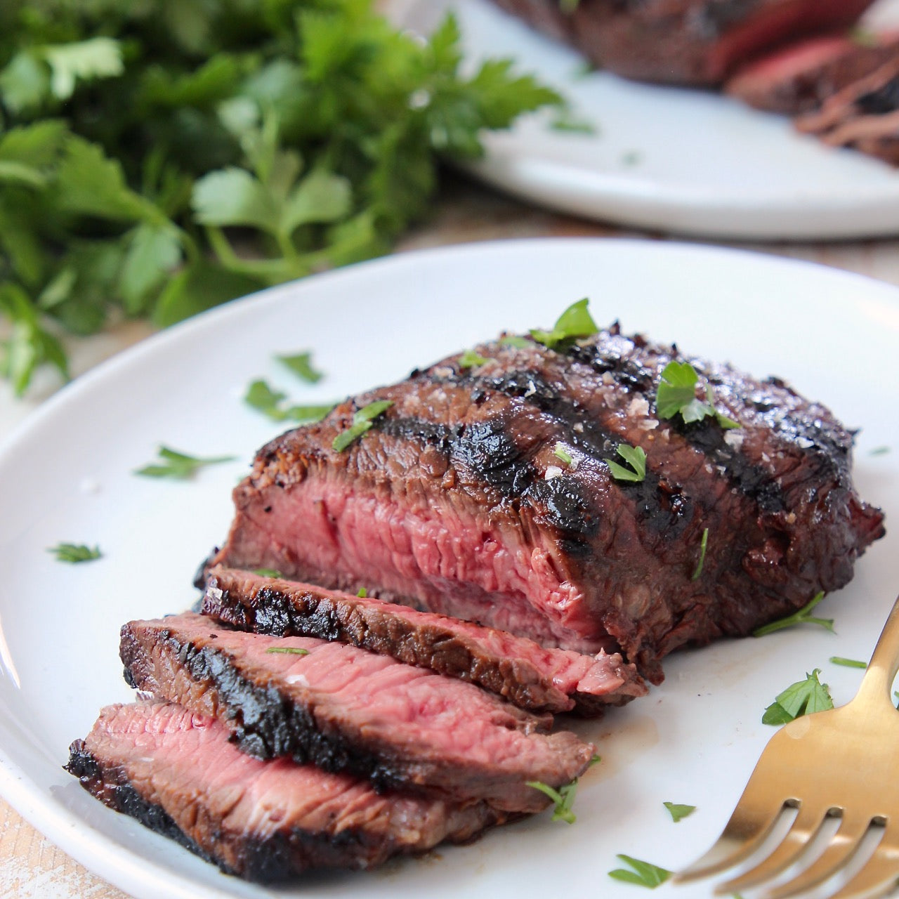 A plate with sliced Sirloin Steak garnished with parsley, accompanied by a gold-colored fork and a bundle of parsley in the background.