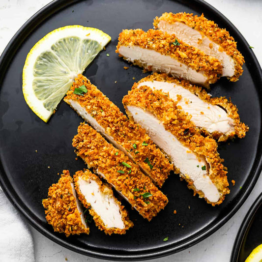 Sliced breaded chicken on a black plate with lemon wedges